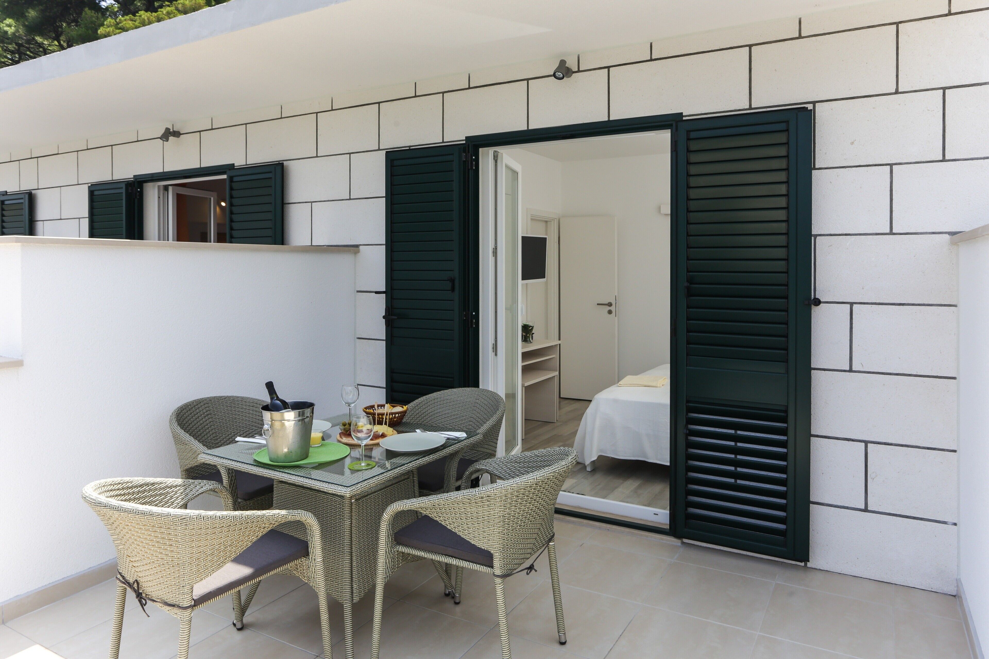 Un espace extérieur aménagé pour les repas, avec des chaises en osier et une table pour deux, un petit coin cuisine équipé d’une cuisinière, et une chambre avec un lit et des volets verts.