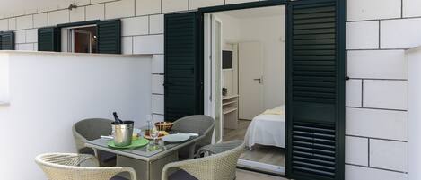 An outdoor dining area with wicker chairs and a table set for two, a small kitchen area with a stove, and a bedroom with a bed and green shutters.