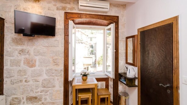 Una habitación con paredes de piedra, un televisor, una mesita con un florero y una puerta.