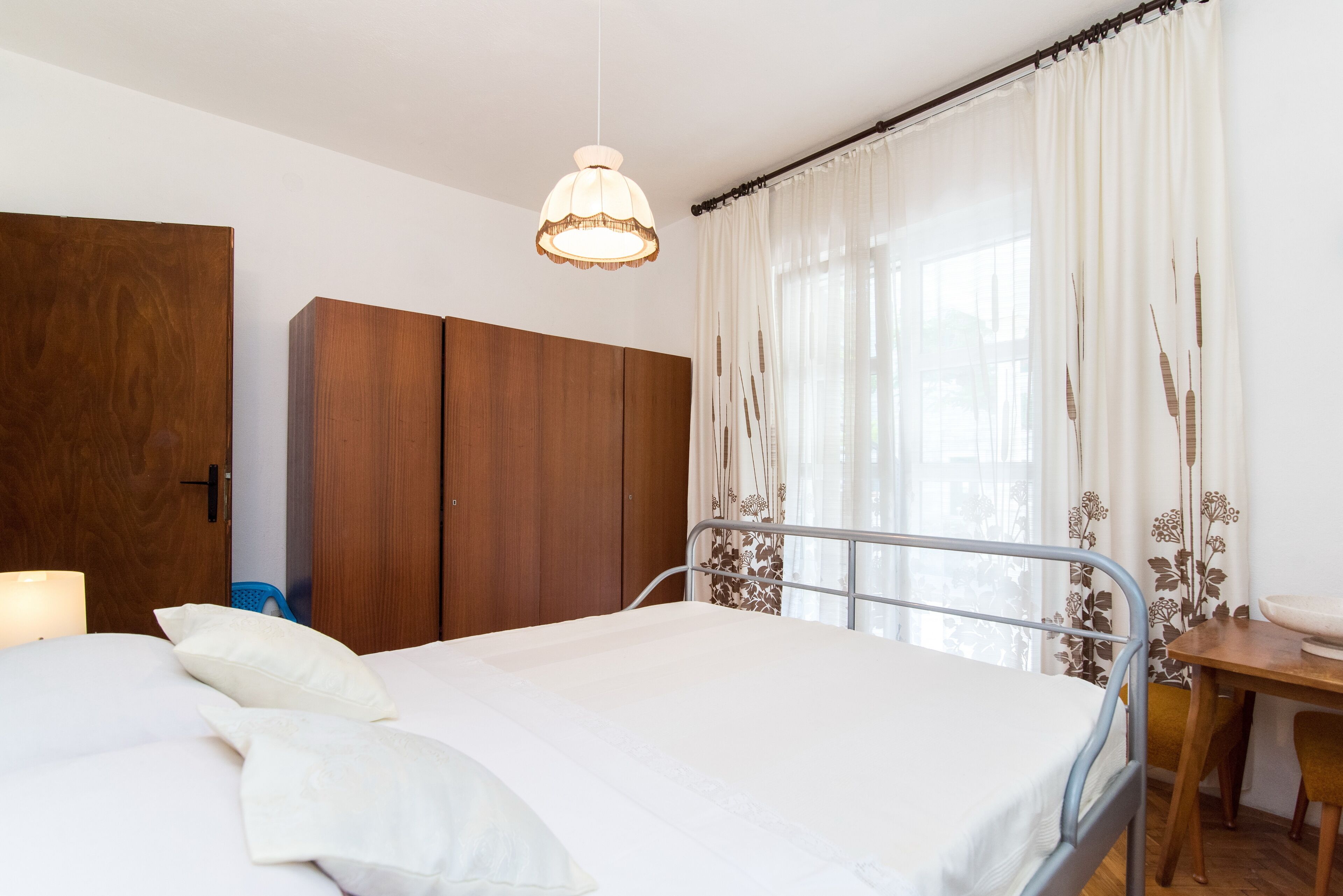 A neatly made bed with white linens, a wooden wardrobe, a metal-framed bed, and a window with sheer curtains.
