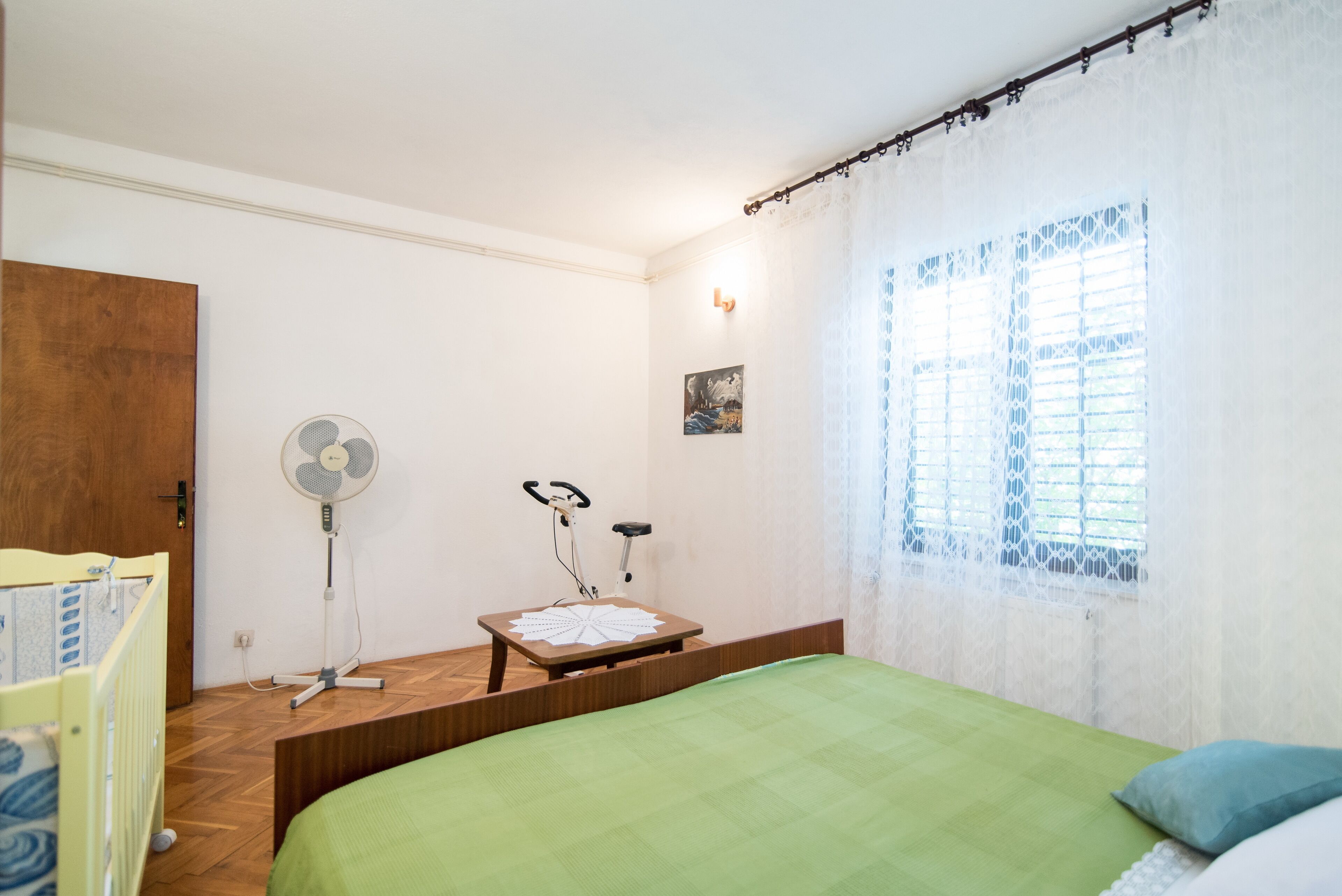Une chambre à coucher avec un lit, un ventilateur sur pied, une petite table, une fenêtre avec des stores et une porte.