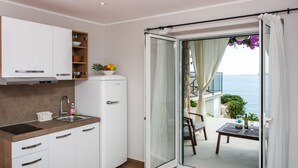 A compact kitchen with a built-in oven, a sink, and a small refrigerator. A sliding glass door leads to an outdoor seating area with a table and chairs, overlooking a body of water.