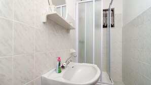 Une salle de bain avec un lavabo blanc, une cabine de douche en verre et une petite étagère au-dessus du lavabo.