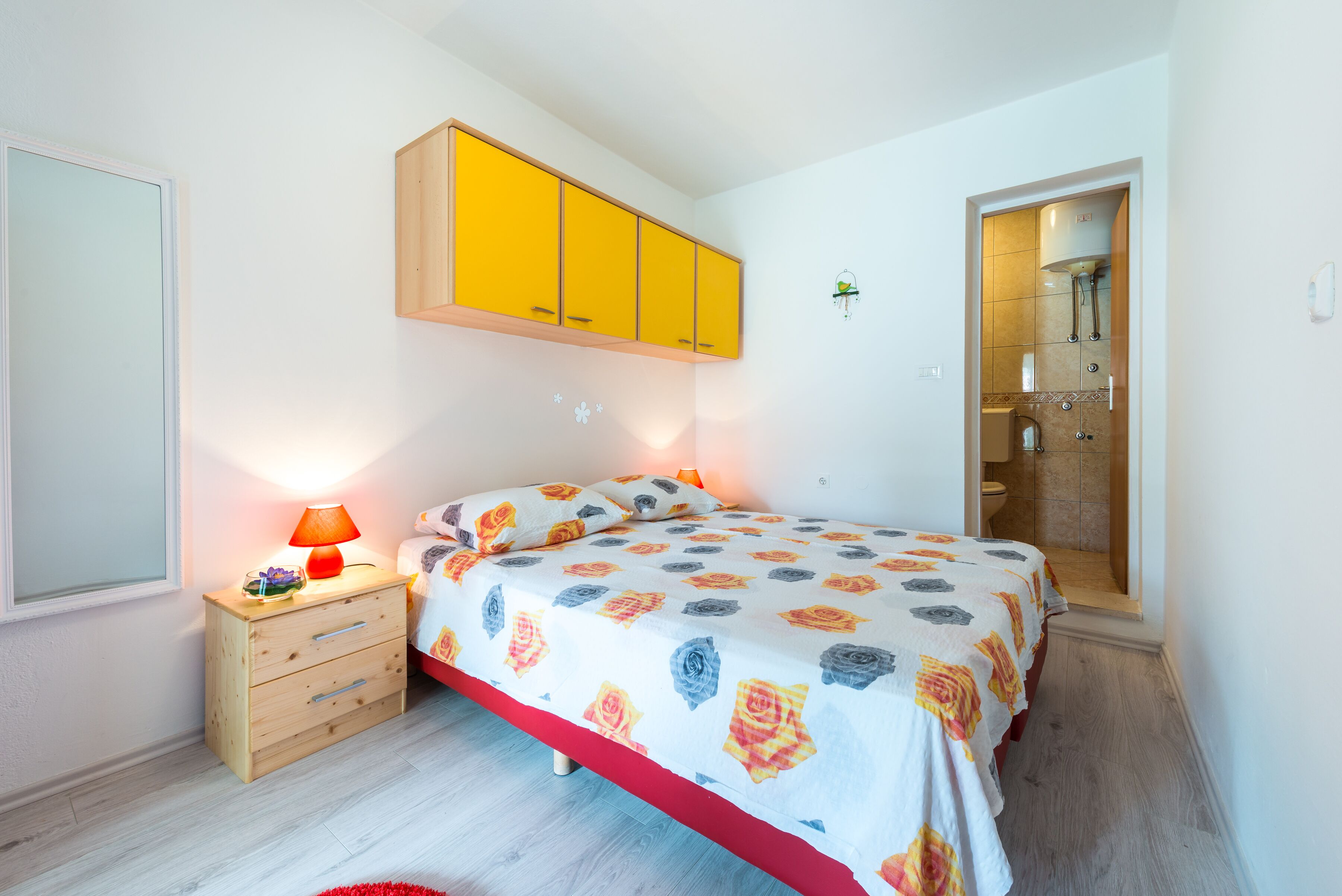 Una camera da letto con un letto, un comodino in legno, una lampada e un bagno visibile attraverso una porta aperta.