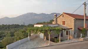 A stone house with a red-tiled roof, a small porch, and a view of mountains in the background.