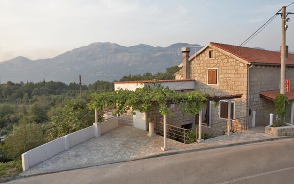 A stone house with a red-tiled roof, a small porch, and a view of mountains in the background.