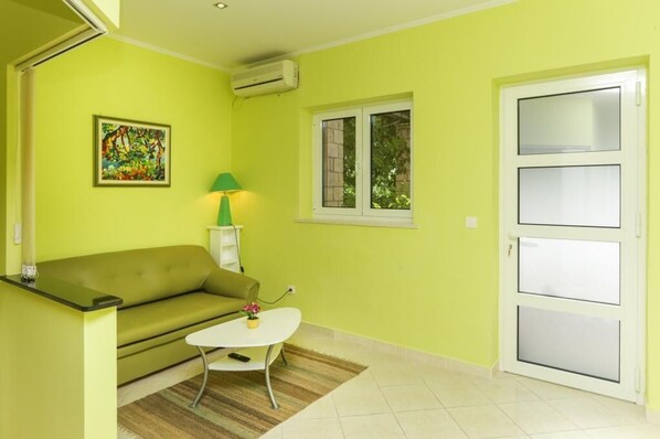 Une pièce avec un canapé vert, une petite table blanche, une lampe verte et une fenêtre donnant sur un paysage verdoyant.