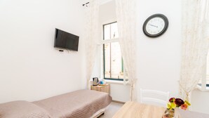 Une petite chambre bien éclairée, avec un lit simple, une table en bois, une horloge et un téléviseur fixé au mur.