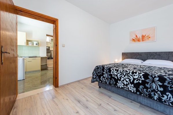 A bedroom with a bed, a wooden door leading to a kitchen, and a wall-mounted artwork.