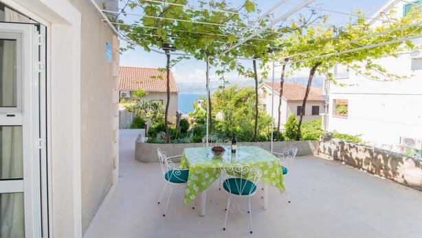 An outdoor patio with a table and chairs, a pergola with vines, and a view of the sea and buildings in the background. - Apartments Veve - Studio Apartment with Terrace Sea View (Postira)
