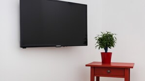 Una habitación con una mesa blanca, un mesita auxiliar roja con una planta, y un televisor negro montado en la pared.