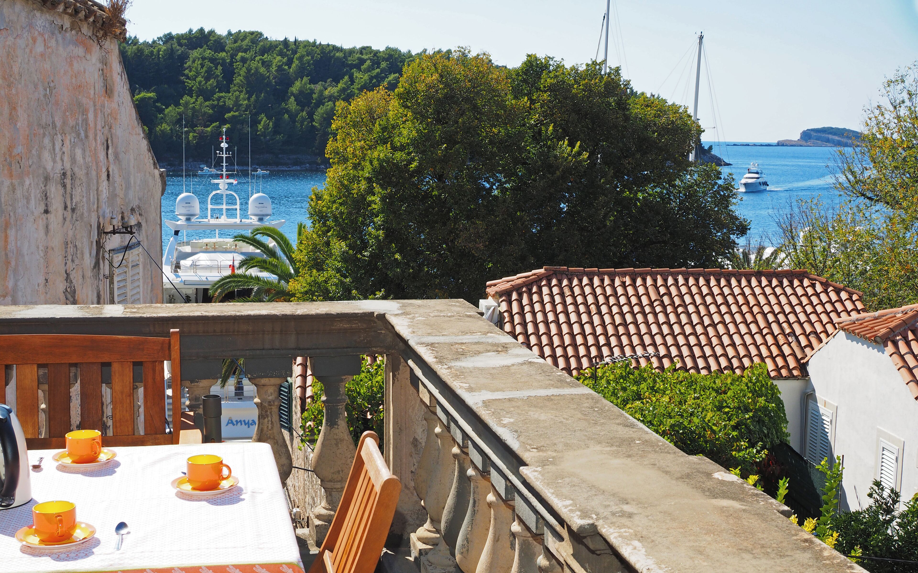 A terrace with a table set for two, overlooking a coastal area with boats and greenery.