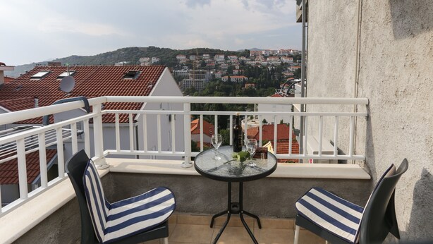 A balcony with two striped chairs, a small round table with two wine glasses and a bottle, and a view of a town with red-roofed buildings.
