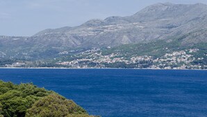 A coastal town with buildings along the shoreline, a mountain range in the background, and a clear blue sea in the foreground.