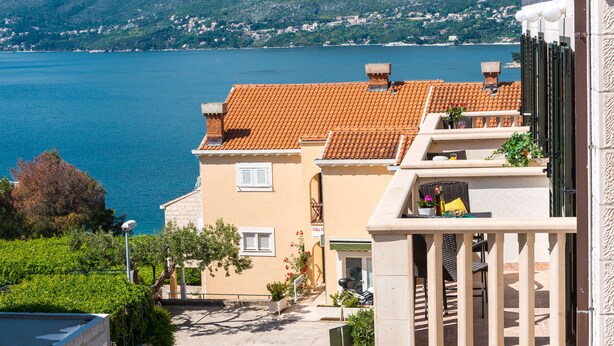 A coastal building with a tiled roof, a balcony with outdoor seating, and a view of the sea and mountains.