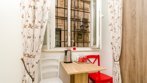 Un petit coin repas avec une table en bois et deux chaises, une blanche et une rouge, près d’une fenêtre ornée de barreaux en métal.