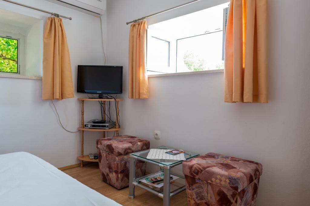 A small room with a bed, a TV on a shelf, two patterned ottomans, a glass table, and curtains.