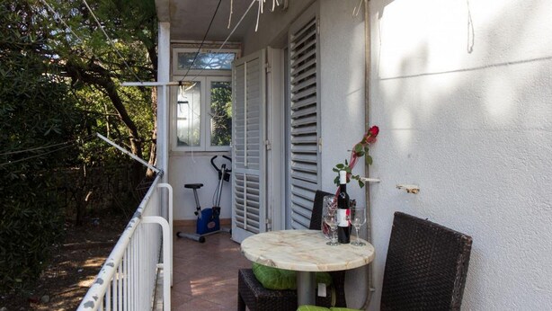Un piccolo patio esterno con un tavolo rotondo, due sedie in vimini, una bottiglia di vino e una rosa sulla parete.