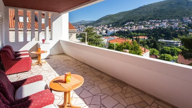 Un balcón con sillas tapizadas de rojo, una mesita redonda y vistas a un pueblo y colinas.