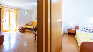 Une pièce avec une porte en bois, un lit recouvert d’une couverture jaune, une table de chevet avec une lampe, un canapé agrémenté d’un coussin à motifs floraux, une petite table avec un bol, et une chaise.