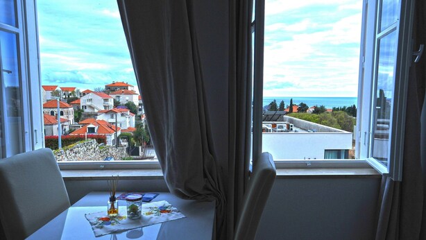 Ein Zimmer mit Tisch und Stühlen, einem fensterbelaubten Fenster und Blick auf Gebäude sowie ein Gewässer.