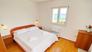 Un dormitorio con piso de madera, una cama con ropa blanca, una mesita de noche, una lámpara de cabecera, un ventanal con vista al mar y un mueble pequeño.