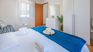 Ein Schlafzimmer mit einem Bett, einem Schminktisch, einem Spiegel, einem Fenster mit Vorhängen und einem kleinen Tisch mit einer Kerze.