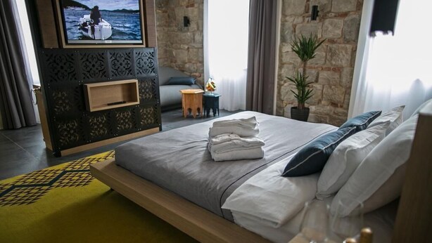 A modern hotel room with a large bed, a flat-screen TV mounted on a decorative wall, a small seating area with a sofa and side table, and a stone wall in the background. - Rooms Lejletul  - Superior Double Room with Square View (Kadr) (Split)