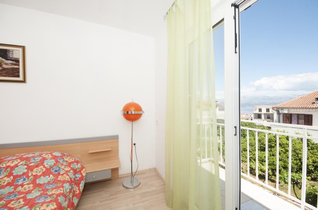 A bedroom with a bed, a floor lamp, a window with curtains, and a balcony with a view of buildings and greenery.