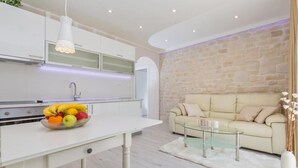 A modern kitchen with white cabinets, a fruit bowl on the counter, and a dining area with a glass table and sofa.