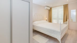 A bedroom with a white bed, a bedside table, a nightstand, and a window with curtains.