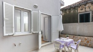 An outdoor patio with a table and chairs, a potted plant, and a building with a stone wall and a window with shutters. - Apartments Villa Karmen - Studio with Terrace (Lavanda) (Dubrovnik)