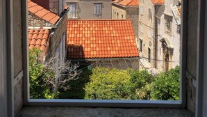 Blick durch ein Fenster auf ein Gebäude mit roten Ziegeldächern, Steinmauern und einer Mischung aus Baustilen, darunter ein Turm und ein Glockenturm.