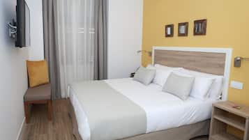A hotel room with a bed, a bedside table, a TV mounted on the wall, and a window with curtains.