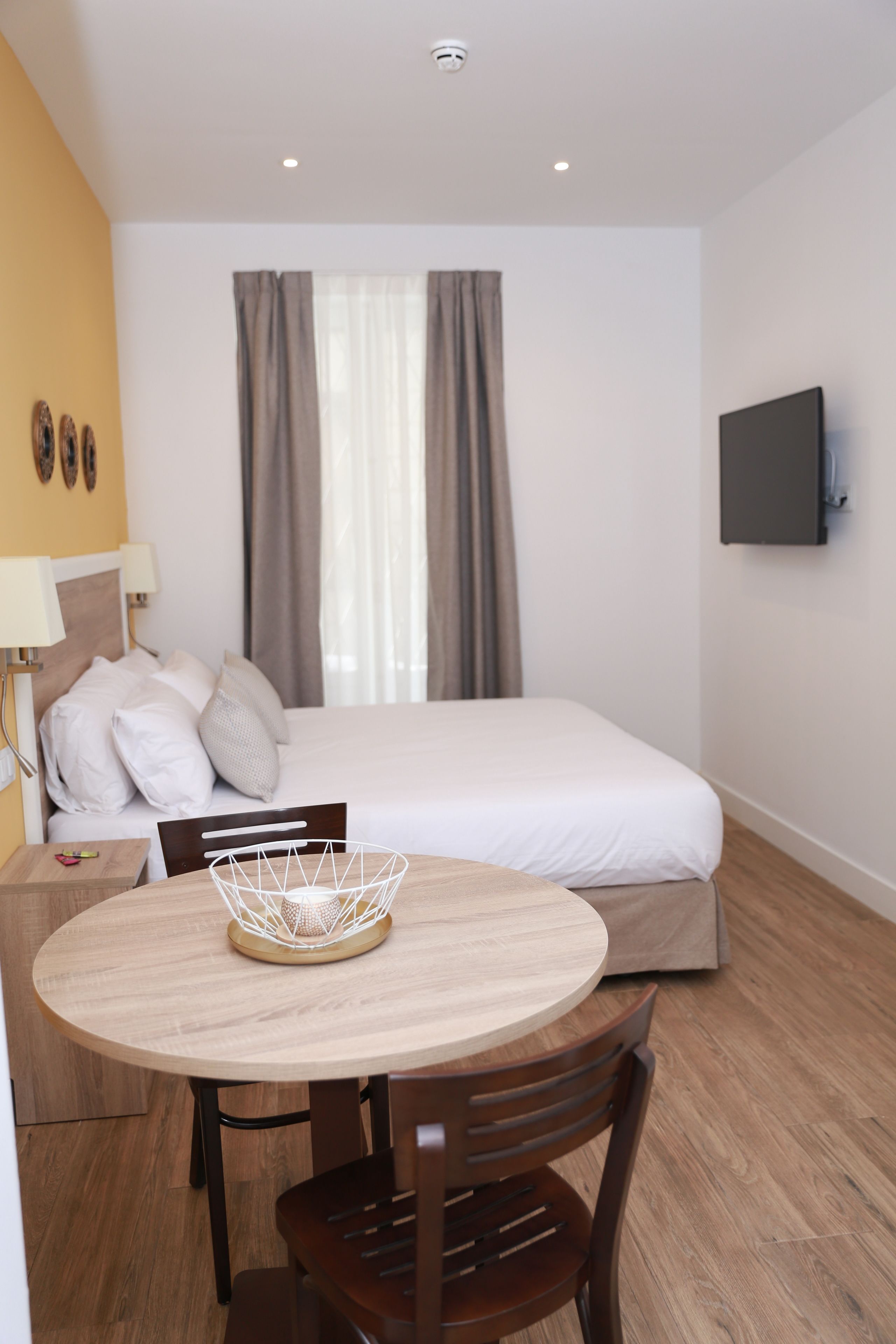 Habitación de hotel con cama, televisor montado en la pared, una mesa de comedor con sillas y una ventana con cortinas.