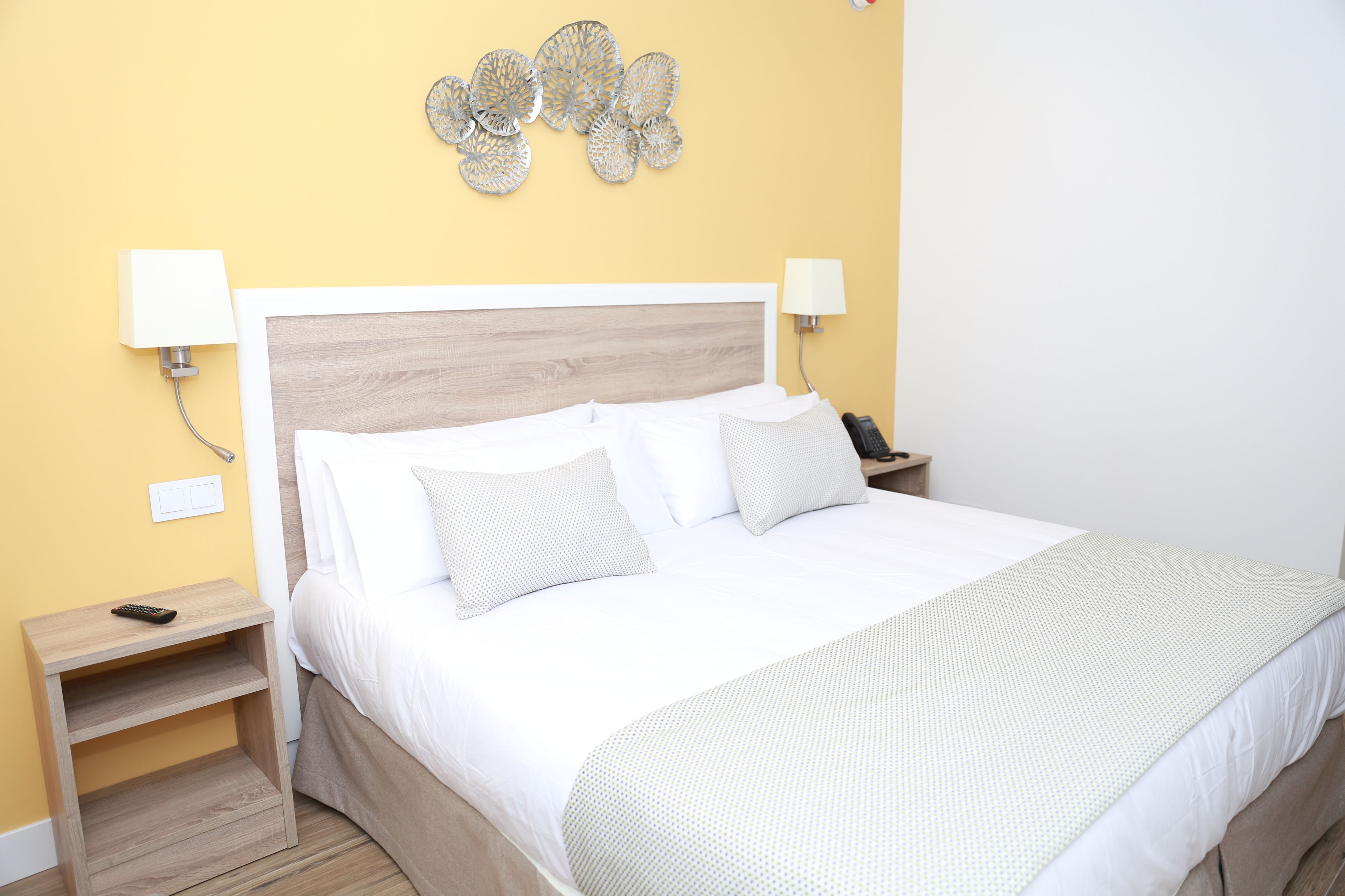 Een netjes opgemaakt bed met witte lakens en een lichtgroene deken, een houten nachtkastje met een afstandsbediening, en een decoratief object aan de muur.