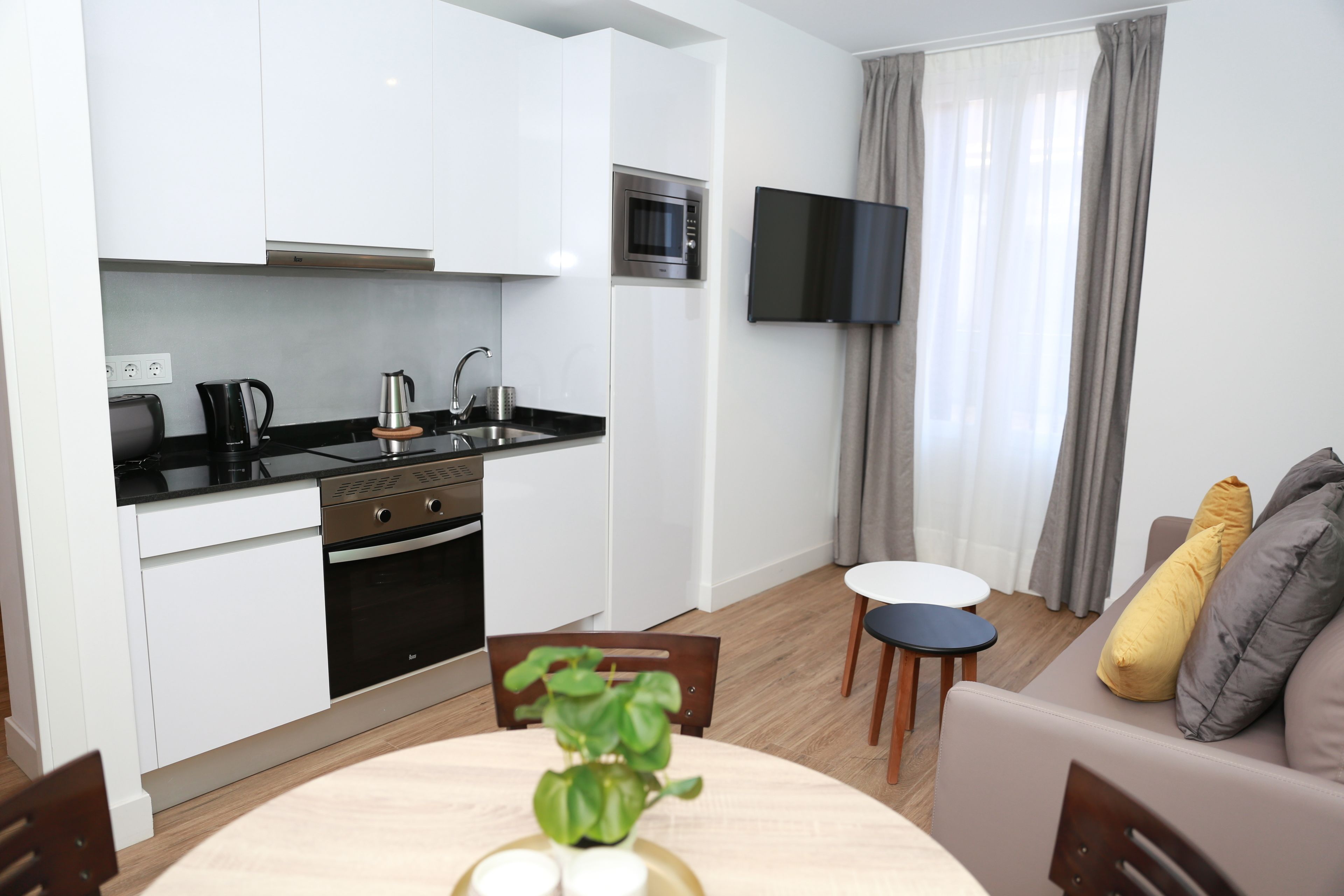 Eine moderne Küche mit weißen Schränken, einer eingebauten Mikrowelle und einem schwarzen Herd. Ein Esstisch mit Stühlen und ein Blumentopf sind vorhanden. Ein Flachbildfernseher ist an der Wand montiert, und ein Sofa mit Kissen ist zu sehen.