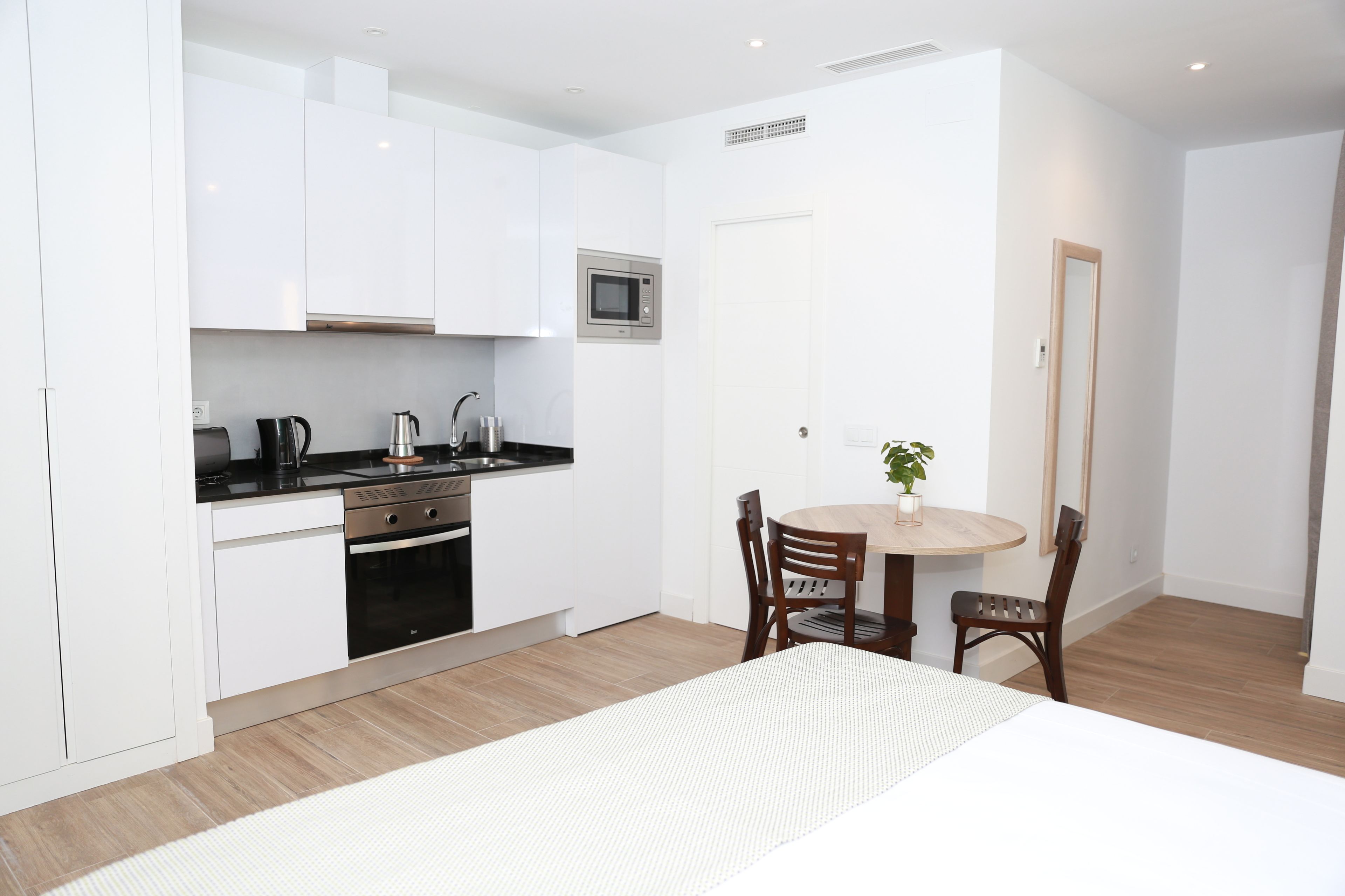 Une cuisine compacte avec des meubles blancs, un four à micro-ondes, une bouilloire et une cuisinière. On y trouve une table à manger avec deux chaises et une petite plante en pot.