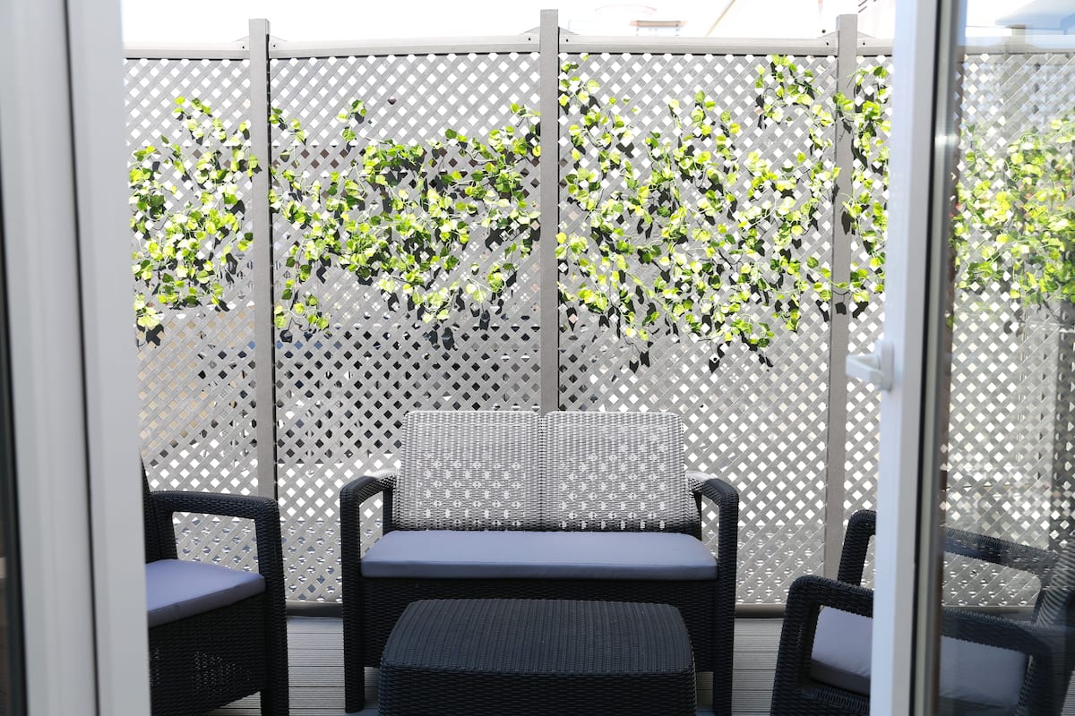 Un espace de détente extérieur aménagé avec des meubles en osier, comprenant un banc rembourré et deux chaises, une clôture en treillis ornée de verdure et une cloison en verre transparent.
