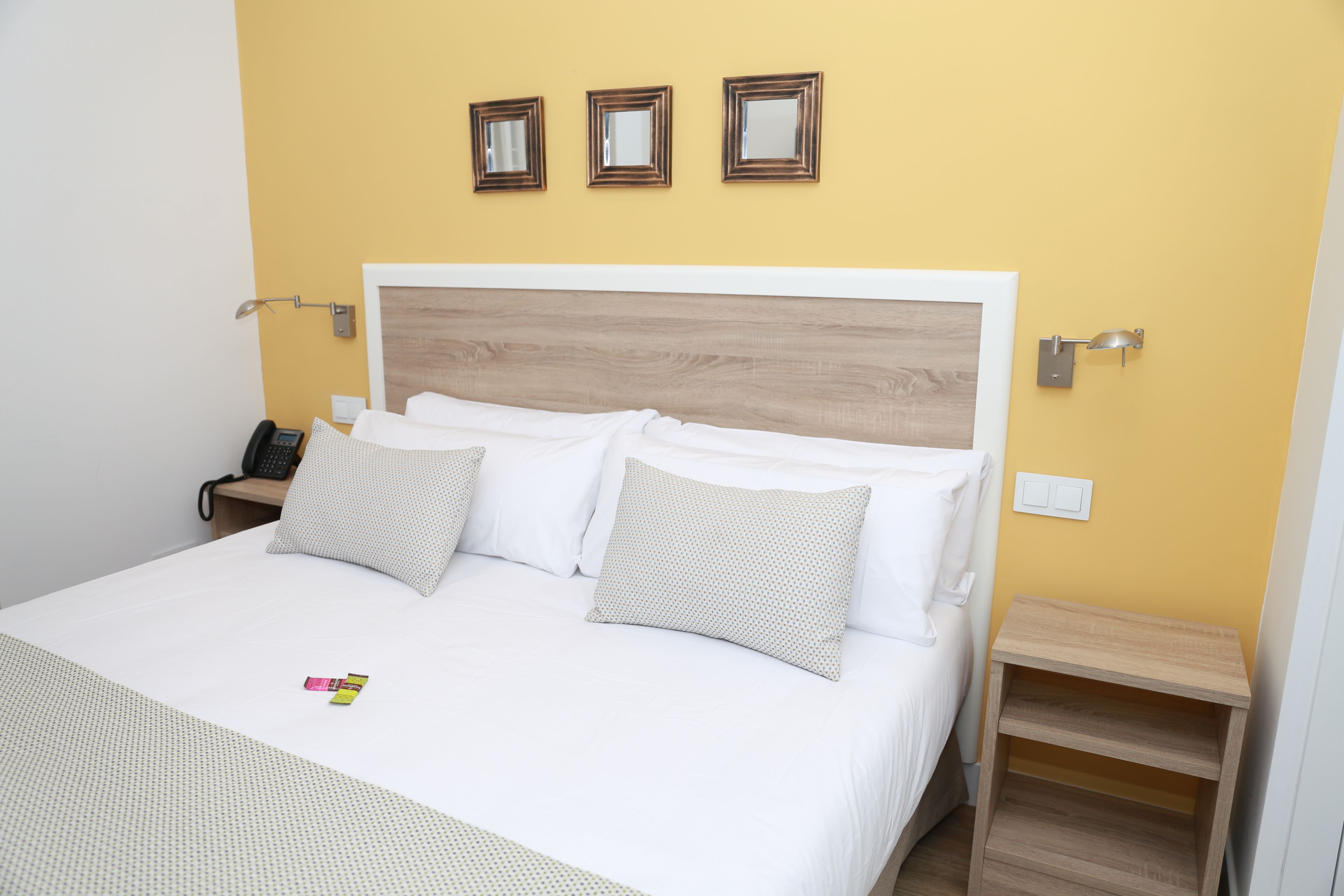 Een netjes opgemaakt bed met witte beddengoed en twee kussens, een nachtkastje met een lamp, en drie inlijstingen met spiegels aan de muur.