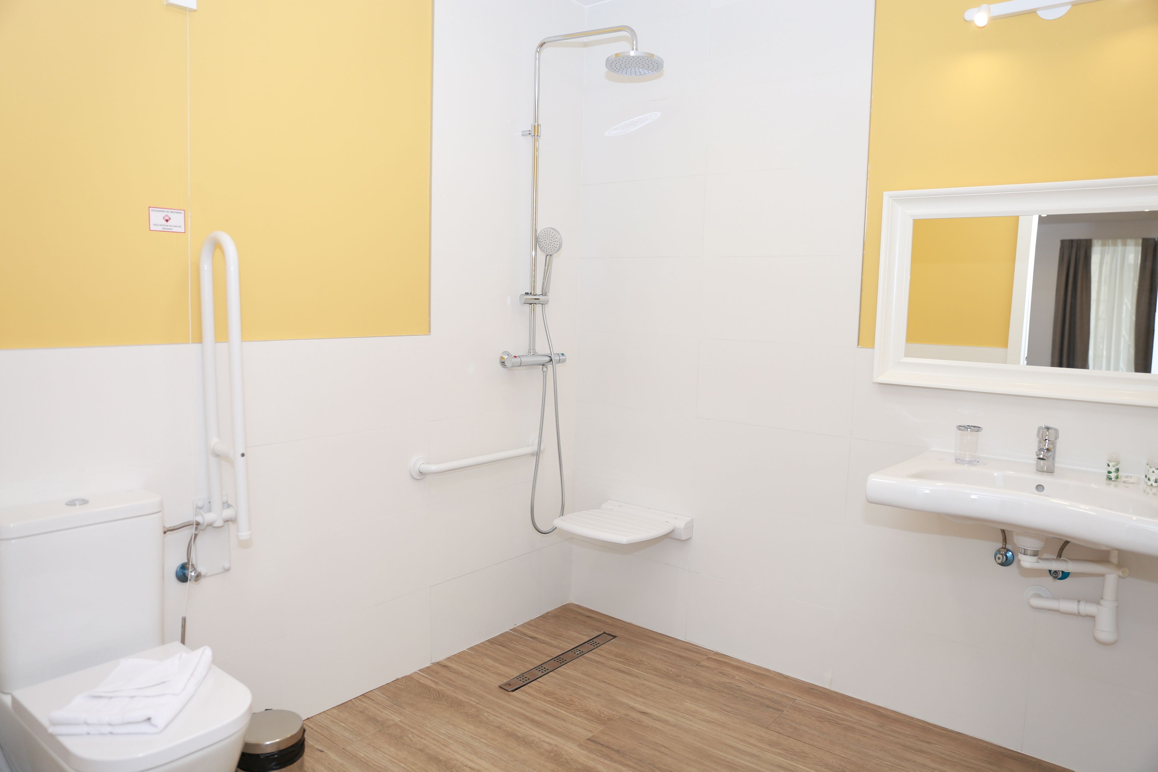 A bathroom with a white toilet, a shower with a handheld showerhead, a sink with a mirror, and a wooden floor.
