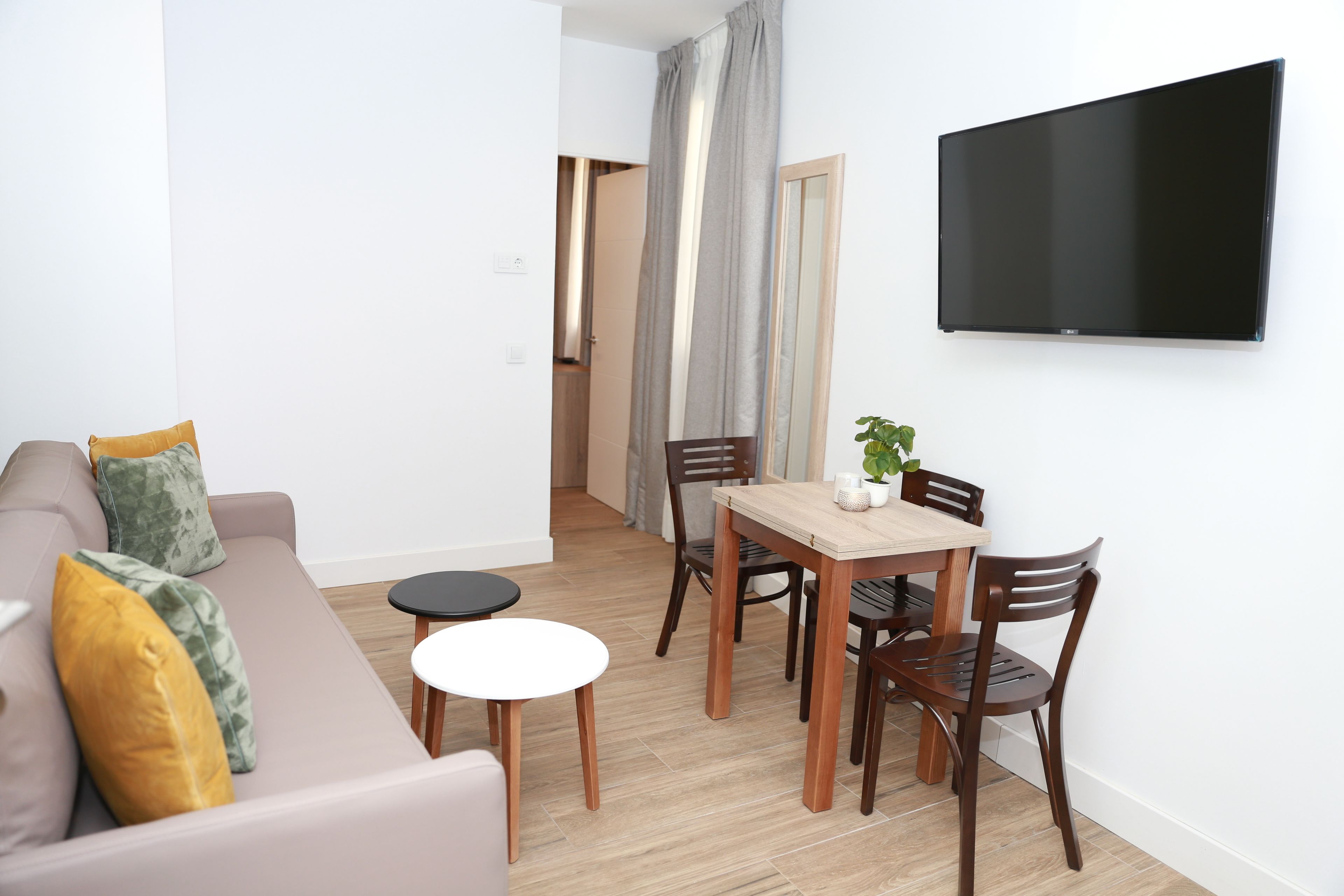A modern living room with a sofa, a dining table with chairs, a small round table, and a flat-screen TV mounted on the wall.