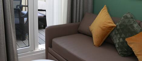 A modern living room with a brown sofa, two cushions, a small white table with a candle, and a view through a glass door to an outdoor area with chairs and greenery.