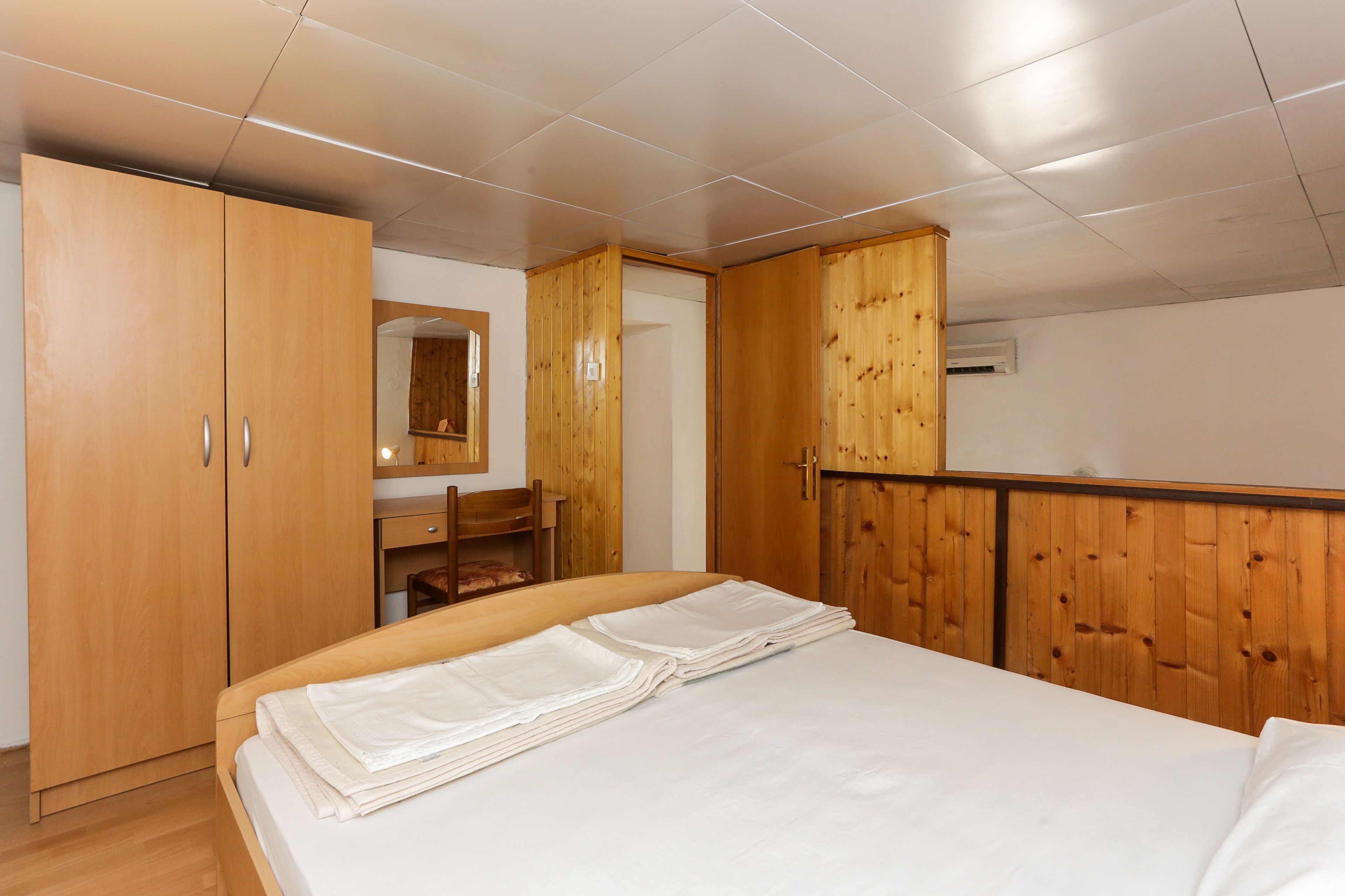 Une chambre avec une armoire en bois, un lit avec du linge de lit blanc, une tête de lit en bois et un miroir au-dessus de la commode.