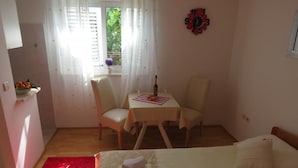Ein Hotelzimmer mit einem Bett, einem kleinen Tisch mit einer Flasche und Gläsern, einem Sessel, einem Fenster mit Vorhängen und einer Wanduhr.