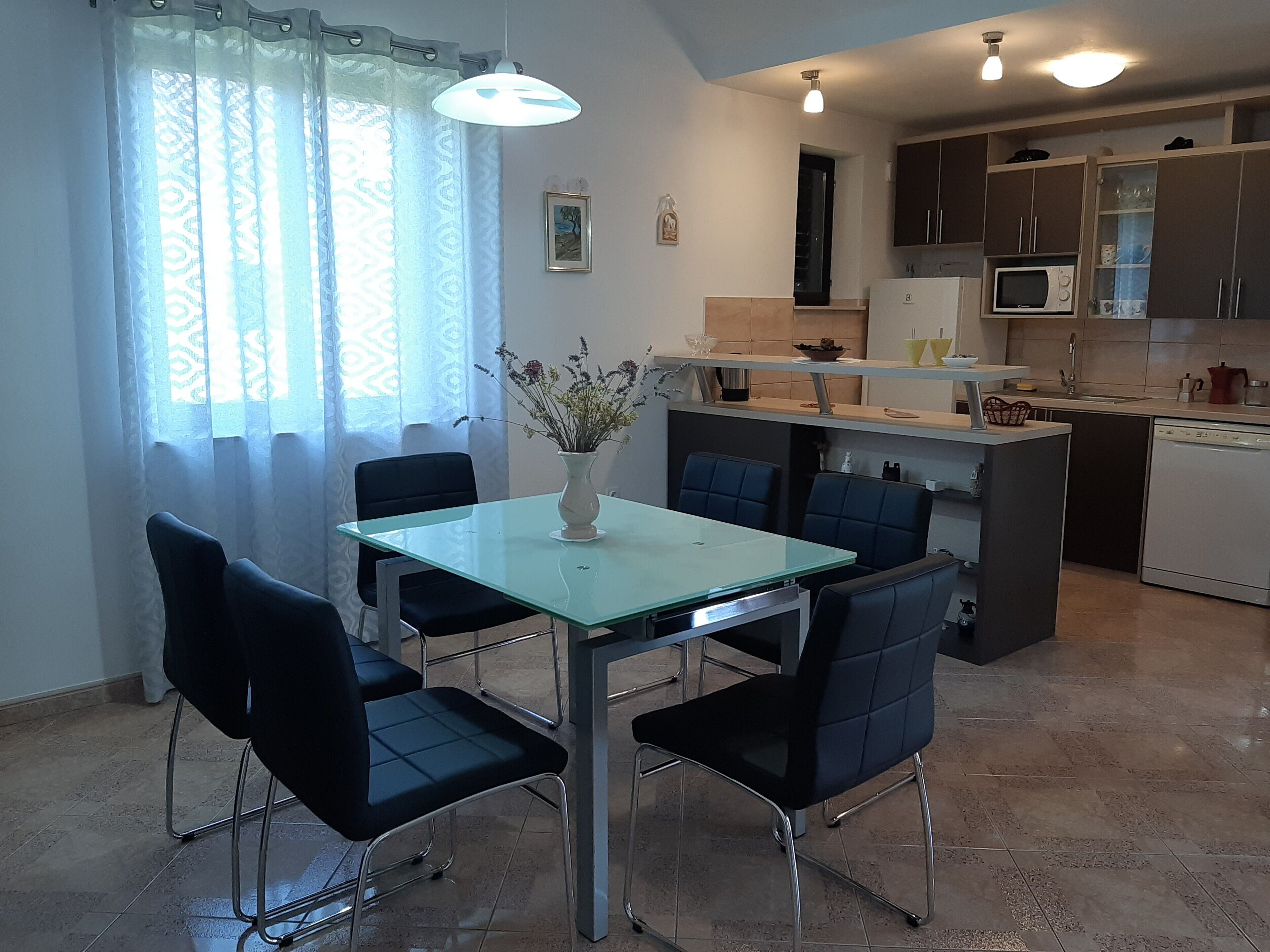 A modern kitchen with a dining area, featuring a glass table, black chairs, and a vase with flowers.