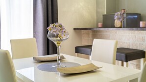 Una mesa de comedor con un florero de vidrio transparente con flores, un mantel individual y una servilleta doblada. Hay sillas alrededor de la mesa, y se ve una chimenea de ladrillo al fondo.