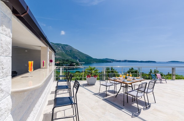 Une terrasse avec une table dressée pour le repas, des chaises, un pot de fleurs et une vue sur la mer et les montagnes.