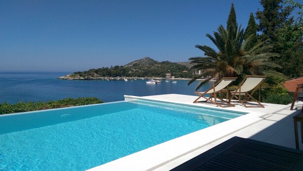A pool with clear blue water, two lounge chairs, and a view of the sea and a hill with buildings in the background.