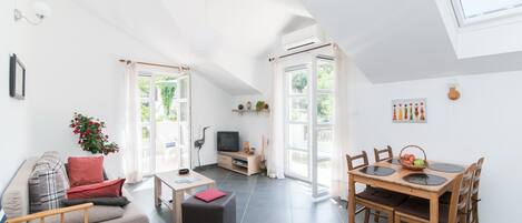 A spacious living area with a sofa, ottoman, and dining table with chairs. There is a television and a window with curtains.